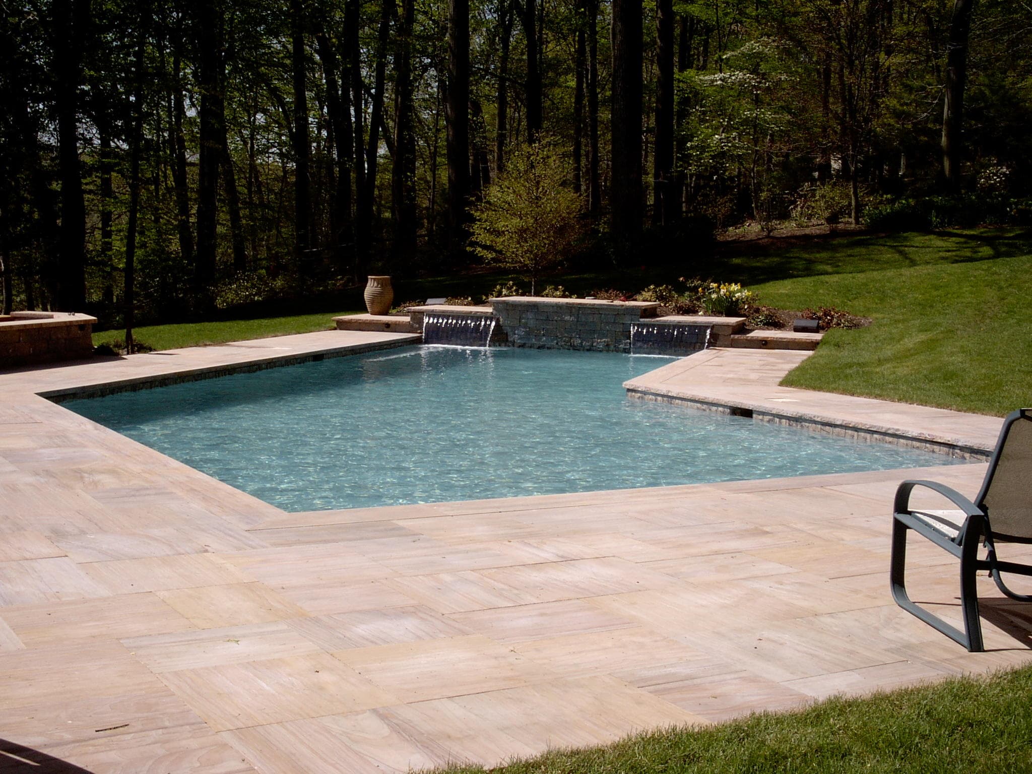 Rectangular pool with sheer descent water feature and travertine deck in wooded backyard
