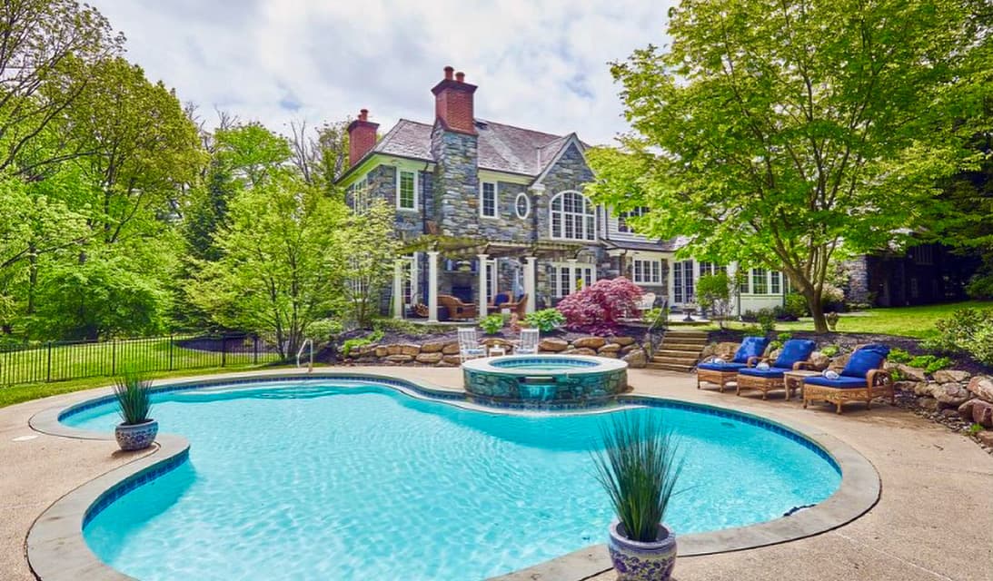 Freeform pool with raised stone spa and bubblers behind stone-front estate home with red maple