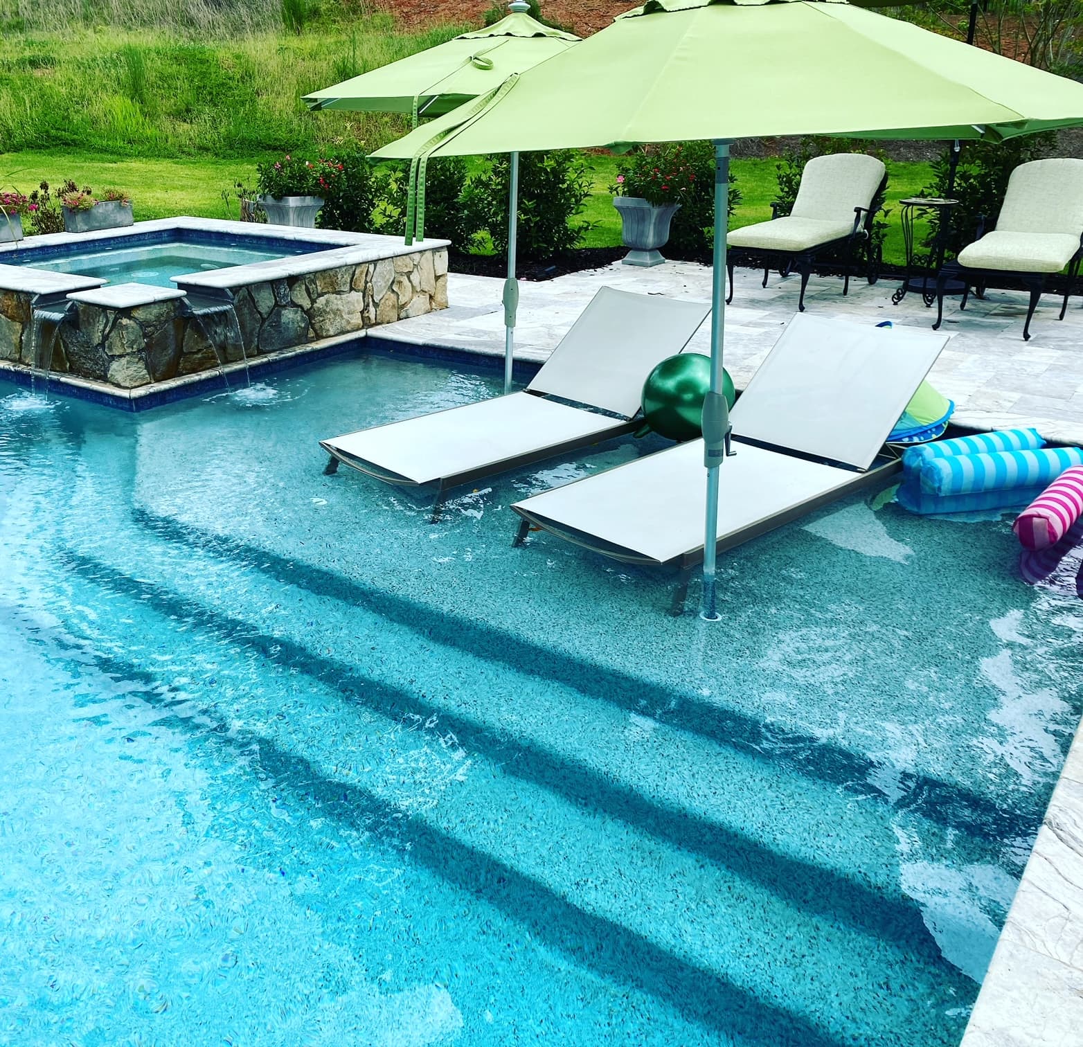 Pool tanning ledge with white in-pool loungers under green umbrella beside stone spa
