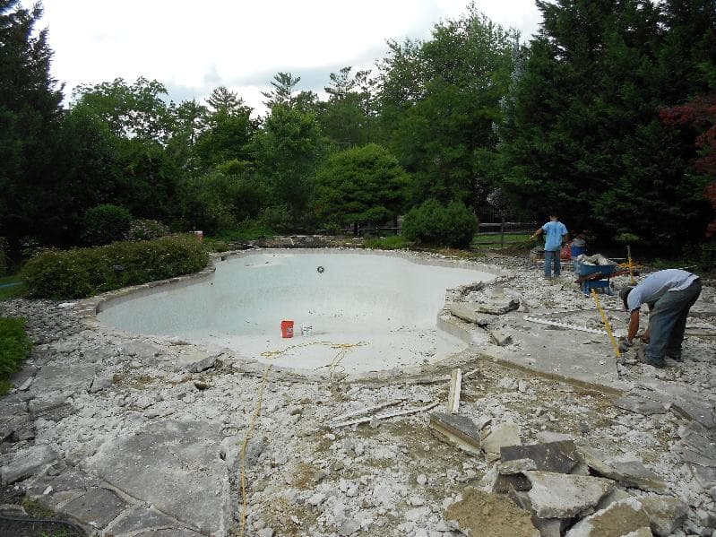 Older freeform pool with green water during deck rebuild with crew at far end