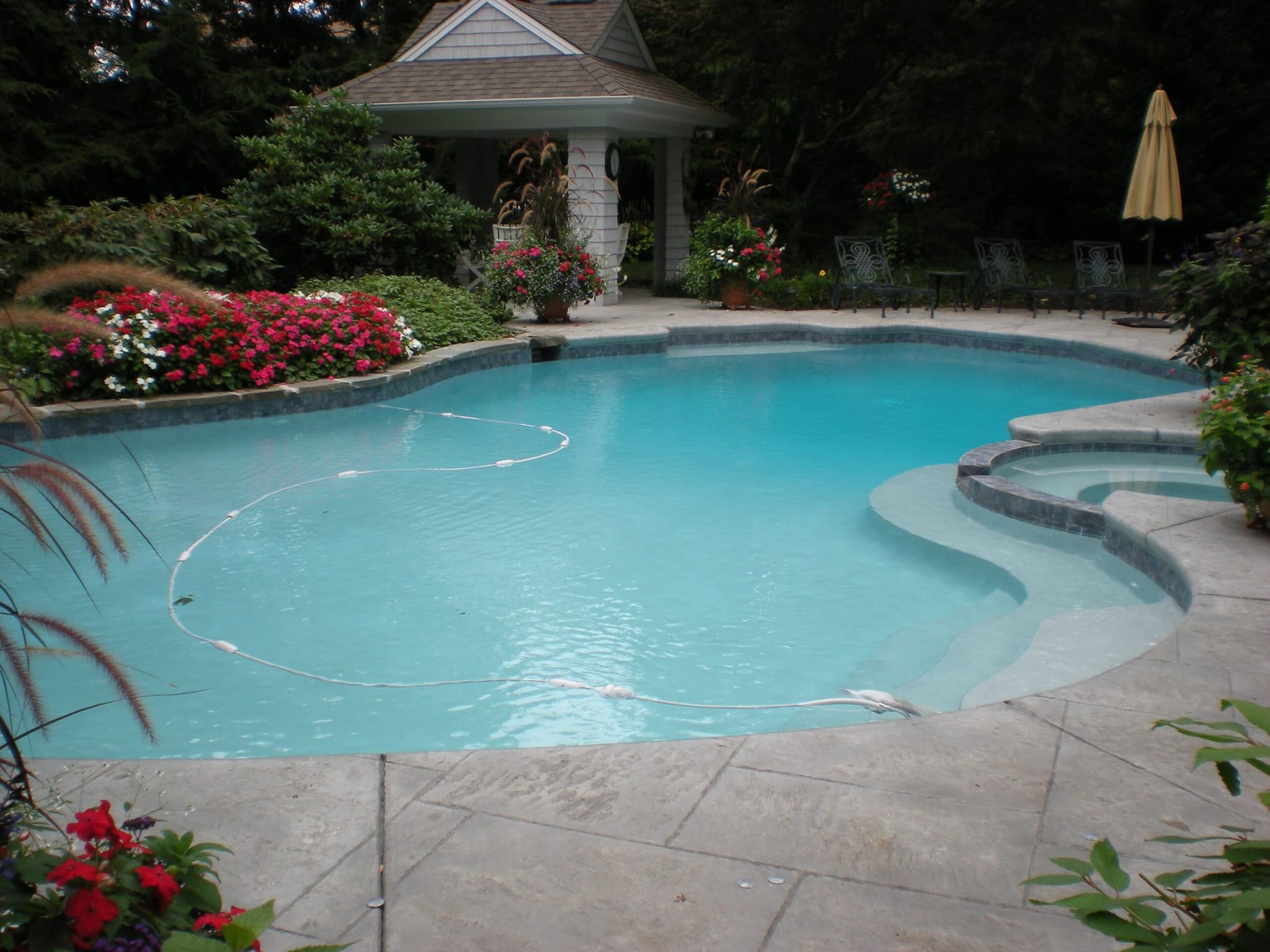 Freeform pool with raised step area and pool house cottage with red flower borders