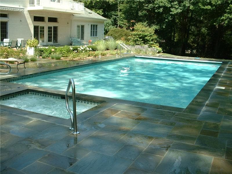 Rectangular pool with attached spa and full bluestone deck behind white brick home
