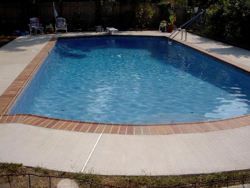 Rectangular pool with red brick coping in fenced backyard with towels on chairs
