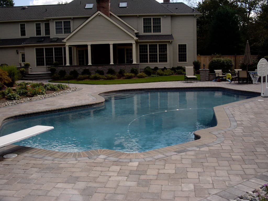Freeform pool with paver deck and diving board behind tan colonial home